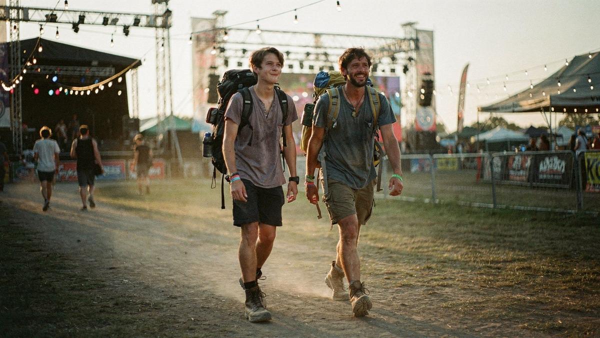 Two young travellers with backpacks walking a dusty path between festival stages at dusk